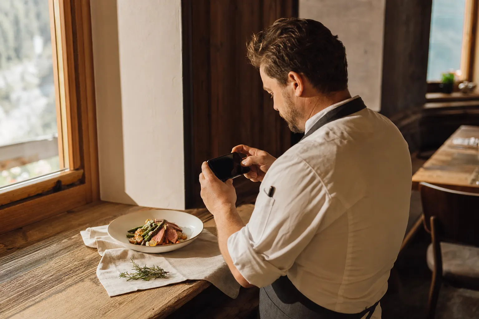 Person in Kochjacke fotografiert einen angerichteten Teller mit dem Smartphone am Fenster